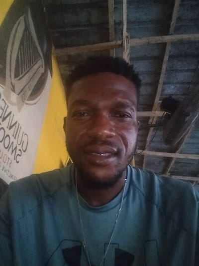 A man in a teal t-shirt and silver chain smiles at the camera. The background is a cluttered space with a Guinness advertisement and a wooden ceiling. A fan hangs from the ceiling. The lighting is dim, creating shadows on the man's face. His hair is short and dark.