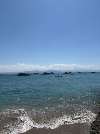 In Kampung Toyapakeh, Indonesia, a serene seascape unfolds under a clear blue sky. The turquoise waters of the ocean, shimmering with sunlight, gently lap against a dark, rocky shore in the foreground. A line of white-capped waves advances, leaving a foamy trail on the sandy beach.
Further out, a fleet of boats dots the horizon. Several larger vessels, likely passenger ferries or tour boats, are anchored or cruising leisurely. Closer to the shore, a smaller, green boat with two figures on board navigates the water. In the distance, a hazy mountain range forms a majestic backdrop, with a scattering of puffy white clouds adorning the sky above.