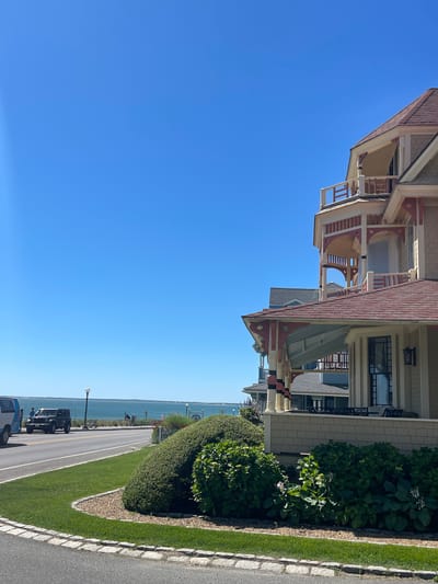 In Oak Bluffs, USA, a bright, clear day unfolds with a view of the ocean. The foreground is dominated by lush green landscaping and the ornate facade of a Victorian-style house, featuring a multi-level balcony with white railings and decorative red accents. Adjacent houses, also exhibiting classic architectural styles, are visible further down the street. A paved road runs parallel to the coastline, where a dark-colored Jeep and a white van are parked. Several individuals are seen standing near the vehicles and along the waterfront promenade, which is lined with trees and lampposts. The tranquil ocean stretches to the horizon under a vast, cloudless blue sky.