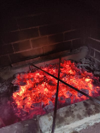 The image depicts a close-up view of glowing red-hot embers in a fireplace. Two dark metal rods are positioned diagonally across the embers, partially obscuring them. The background consists of dark brickwork, suggesting the interior of a hearth. The overall lighting is dim, with the primary illumination coming from the intense heat of the coals. There are no people or animals visible in the scene. The environment appears to be indoors, likely a residential setting in Востец Снт, Russia. The image captures a moment of intense heat and potential activity related to a fire.