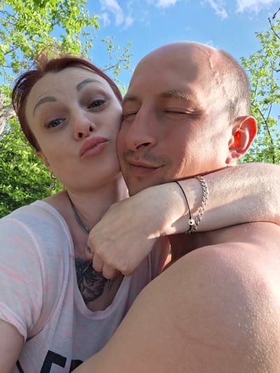 The image shows a man and a woman lying in an outdoor setting, likely a park or yard, in Сваткаўскі сельскі Савет, Belarus. The woman, with auburn hair pulled back, a fair complexion, and distinct eyebrows, is in the foreground with her arm draped around the man's neck. She is wearing a light-colored shirt and a silver chain. She is pouting her lips toward the camera. The man has a shaved head and closed eyes. He is bare-chested. Both are facing upwards toward the sky visible through tree branches. The woman has a tattoo on her chest. The man has a bracelet on his wrist.