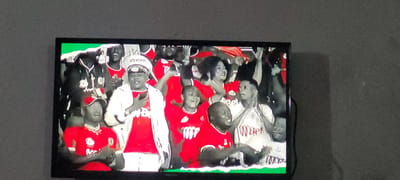 The image displays a television screen showing a crowd of people, presumably at a sporting event or concert, given the vibrant atmosphere and red attire worn by many individuals. The people appear to be cheering and enjoying themselves, with some captured in moments of laughter and excitement. One prominent figure in the center wears a white captain's hat and a red shirt, holding a phone. Another person in a red shirt is visible on the left side of the screen wearing a red baseball cap. The overall scene suggests a lively and engaged audience. The television itself is positioned on a wall in a room, with the screen taking up a significant portion of the frame. The wall behind the television is a textured, dark gray, possibly concrete or plaster. The lighting in the room appears to be dim, with the primary illumination coming from the television screen. The television has a black bezel and is mounted or placed on a surface that is not fully visible. There is a hint of green at the bottom edge of the television screen, suggesting a graphic overlay from the broadcast. This image was captured in Kipala Mpakani, Tanzania.