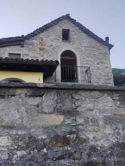 The image shows a stone building with a tiled roof, likely a church or a historical structure. In the foreground, a rough-hewn stone wall dominates the lower half of the frame. Above this wall, a portion of the building is visible, constructed from lighter-colored stone. The building features a small window, an arched doorway leading to a balcony with an ornate metal railing, and a decorative stone relief above the doorway. A portion of a yellow wall is visible beneath a dark overhang with terracotta tiles. The background shows a light sky and some greenery on the right side. The building appears to be located in the town of Orselina, Switzerland, given the architectural style and the surrounding landscape which is typical of the region.