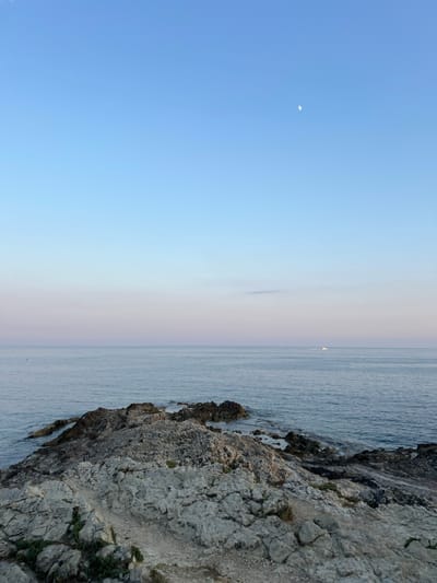 A seascape featuring a rocky coastline on the right. The water stretches towards the horizon, blending with a pale sky. In the upper left, a small crescent moon is visible in a clear blue sky. A distant white sail is visible on the water. The transition from the light blue sky to the sea shows a gradient of colors. The rocks are grey and rugged, with sparse patches of green vegetation.
