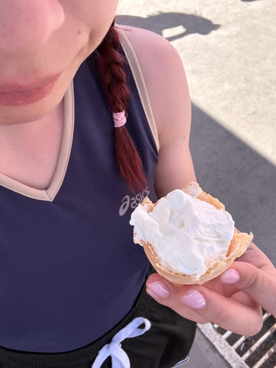 A woman is lying down outside in Bekhterevo, Russia, holding an ice cream cone in her right hand. The cone is mostly eaten, with only a bit of ice cream remaining. She is wearing a dark blue athletic top, with a cream-colored neckline, black shorts with a white drawstring, and her hair is in a red braid secured with a pink hair tie. Her nails are light pink with glitter. The background is concrete and a grated metal surface.