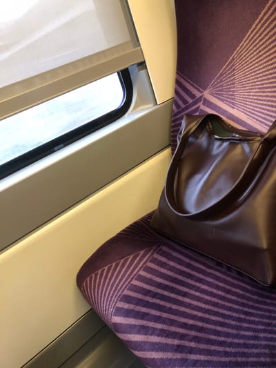 Within a train in Bernolsheim, France, a brown tote bag rests on a purple upholstered seat. The seat features a geometric pattern of radiating lighter purple stripes on a darker purple background. Adjacent to the seat is a window with a black frame, partially obscured by a white roller blind positioned in the upper portion. The window looks out onto a brightly lit, indistinct exterior. The interior paneling surrounding the window and seat is a light beige or cream color. No individuals are present in the frame.