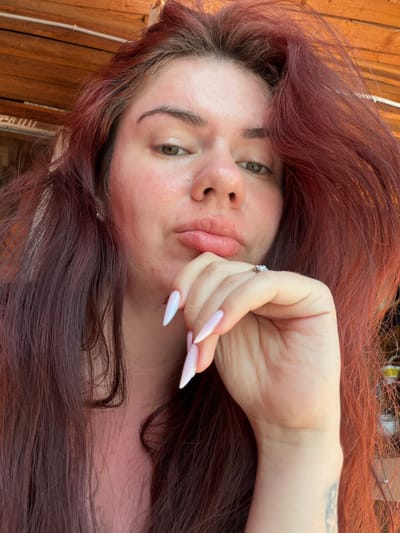 A selfie shows a person with auburn hair and light skin, making a kissing face. They are wearing a ring on their right hand, which is resting on their chin. The background appears to be outdoors, with wooden slats and some foliage. The image is a close-up, focusing on the person's face and upper body. No specific location in Karachino, Russia, can be determined from the image.