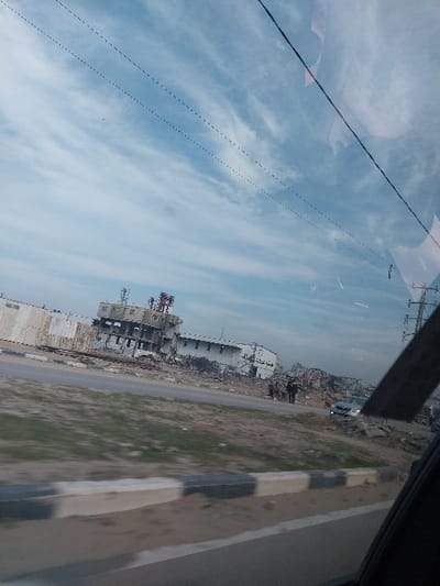 The image depicts a scene of extensive structural damage in a-Zawayda, Palestinian Territory, viewed from the interior of a moving vehicle. In the immediate foreground, the bottom right shows a dark structural element of the vehicle, likely a window frame or pillar. A multi-lane paved road runs horizontally across the mid-ground, featuring a concrete median barrier painted with alternating black and white stripes. Beyond the road, on a slightly elevated area, lies a landscape dominated by severe destruction.

The central subject is a large, multi-story building appearing significantly damaged or partially destroyed, located on the far side of the road to the left. Its facade is severely compromised, revealing internal structures, exposed rebar, and large sections of debris piled around its base. On the roof of this building, multiple communication towers or industrial structures, some featuring red and white elements, are visible. Arabic script is faintly discernable on the remaining walls of the building. To the right of this primary structure, extending along the roadside, are further scattered rubble and remnants of other destroyed buildings.

In the mid-ground, near the right side of the road and amidst the debris, several indistinct human figures, approximately three to four, are standing. A light-colored sedan or hatchback is partially visible near these figures. The ground between the road and the damaged structures consists of dry grass and dirt. Overhead, the sky is light blue with scattered white, wispy clouds, and multiple dark power lines or communication cables traverse the upper portion of the frame. The overall scene suggests a post-conflict or post-disaster environment.