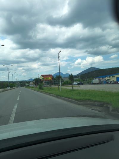 The image shows a road in Goryachevodsky, Russia, with a wide asphalt road extending into the distance. The road is lined with streetlights on both sides. On the right side of the road, there is a grassy area with a white fence and a sign that reads "СВЕТОФОР" (Svetofor) with an arrow pointing to the right and the word "сюда" (syuda) meaning "here". Behind the fence, there are several buildings, including a blue and yellow industrial building, and parked vehicles. In the background, there are rolling hills covered in trees, and a mountain with a telecommunications tower on its peak. The sky is cloudy, with patches of blue sky visible. The image is taken from inside a vehicle, looking out through the windshield.