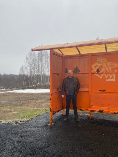A middle-aged man is standing inside a rusted orange structure with a translucent yellow roof. He is wearing a brown leather jacket, a dark shirt, and dark pants. The structure is located in a landscape with patches of snow and bare trees in the background. The ground is a dark, paved surface. The city is Буль-Кайпаново, Russia. There is graffiti on the orange structure behind the man.