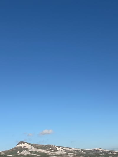 The image shows a steep mountain slope with patches of snow against a clear blue sky. The mountain is rocky and appears to be partially covered in vegetation, with varying shades of brown and green. The snow adds a stark contrast to the earth tones of the mountain. The sky is a vibrant, cloudless blue, suggesting a bright and sunny day. There are no people or objects visible in the scene, and no text is present. The perspective of the image, with the sky occupying the majority of the frame and the mountain running vertically along the right edge, suggests a high vantage point or a wide-angle shot emphasizing the vastness of the sky and the imposing nature of the mountain. The overall impression is one of natural beauty and a remote, possibly mountainous landscape. Given the location context of Jermuk, Armenia, it's likely a scenic view of the Armenian highlands.