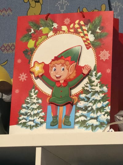 A red Christmas gift bag is displayed on a white shelf in front of a blue, textured wall with a cartoon character. The gift bag features a large circular cutout with an illustration of a cheerful elf wearing a green outfit and a red hat. The elf is holding a star-shaped cookie and is sitting on a blue gift box. Snow-covered Christmas trees flank the elf. The bag is adorned with festive Christmas motifs, including holly, pine branches, and snowflakes. The gift bag's handles are red and red ribbons are visible at the top. The shelf appears to be made of white wood. The lighting suggests an indoor setting, possibly a home or a store. There is no visible indication of Parma, Italy.