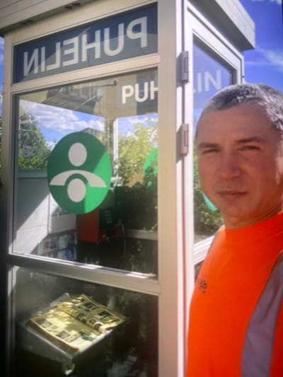The image depicts a man in an orange vest standing next to a public telephone booth in Kerava, Finland. The telephone booth has the word "PUHELIN" (Finnish for telephone) visible on its exterior. Inside the booth, a green circular logo with two white figures is displayed on the glass, and a red telephone unit is visible. A newspaper is placed on a shelf inside the booth. The man is looking towards the camera, and the background shows trees and a blue sky, suggesting an outdoor setting. The overall scene appears to be a street or public area.