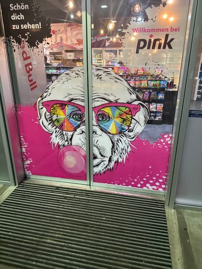 The image displays the entrance to a commercial establishment named "pink" located in Weng im Innkreis, Austria. Two glass doors, set within a metal frame, feature a large, colorful graphic. The graphic depicts a stylized, black-and-white monkey head wearing pink-framed sunglasses with kaleidoscopic, multi-colored lenses. The monkey is shown blowing a pink bubblegum bubble on the lower left door panel. The graphic's background is a vibrant magenta with white splatter effects.

Text overlaid on the glass includes "Schön dich zu sehen!" and "Red Bull" vertically on the left door, and "Willkommen bei pink" with fainter "so cool" and "STORE" below it on the right door. Through the clear sections of the glass, the interior of the store is visible, revealing shelves stocked with numerous brightly packaged goods, likely confectionery or snacks. The store's interior is illuminated by ceiling lights, including decorative exposed-bulb fixtures. In the immediate foreground, a dark grey, horizontally ribbed doormat or floor grating spans the width of the entrance. No individuals are present in the depicted scene.