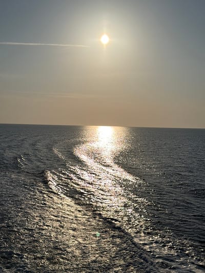 The image depicts an open expanse of sea under a clear sky, observed from the vicinity of Palaiopoli, Greece. The sun, appearing as a brilliant, starburst-like orb, is positioned in the upper-right quadrant of the sky. A faint, horizontal contrail is visible in the upper-left sky. A strong, shimmering golden-white path of sunlight reflects diagonally across the dark water surface, extending from the sun's position towards the lower-left foreground. The water exhibits numerous ripples and distinct wave patterns, with a prominent foamy white wake visible in the lower right, indicating recent passage or presence of a vessel. No landforms or human figures are present.