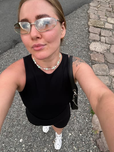 A woman takes a selfie in an outdoor environment. The location appears to be a walkway or path, with stone paving along one side and a gray, gravel-like surface making up the majority of the ground. She is wearing a black dress, a beaded necklace, and white sneakers. She has glasses on and is wearing a black belt-bag. There is a tattoo visible on her left arm. Based on the environmental details and lack of specific landmarks, definitive identification of the exact spot in Jinhua, China, is not possible.
