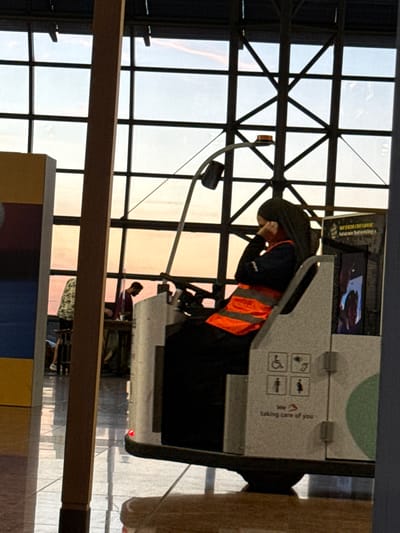 The image captures a scene inside Brussels Airport in Zaventem, Belgium. In the foreground, a person wearing a hijab and an orange high-visibility vest sits in a small, white vehicle, appearing to be on a break, with their hand near their ear. The vehicle has symbols for accessibility and gender-neutral restrooms. Behind them, through a large window with a grid of dark metal frames, a sunset with soft orange and pink hues is visible. The sky is a pale blue. Two other individuals are partially visible in the background through the window, one appearing to be standing near a table. The floor is reflective, showing the ambient lighting of the airport terminal.