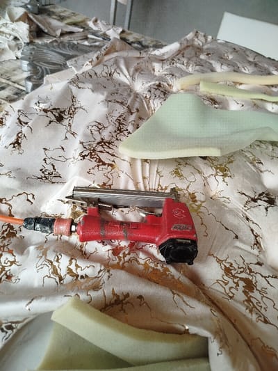 The image captures an indoor scene depicting materials and tools commonly used in upholstery or crafting. At the center and slightly to the left, a red pneumatic stapler or nail gun lies on a patterned fabric. An orange air hose is connected to the back of the tool, with the connector showing some wear and a dark, possibly rubberized, section. The stapler itself is red with a silver-colored magazine where staples would be loaded, and faint lettering on its side appears to spell "BOSTITCH". The tool exhibits signs of use, with some dust or dirt visible on its surface.
The fabric beneath the tool and extending throughout much of the frame is a light, possibly off-white or cream, material with a distinctive metallic gold or bronzed pattern that resembles abstract veins or cracks, giving it a marbled or distressed appearance. The fabric is draped and crinkled in places, suggesting it's actively being worked on or has just been moved.
Scattered across the fabric, particularly to the right and lower right, are several pieces of foam. These foam pieces vary in shape and color, ranging from a light, pale green to a slightly yellowish-green hue. One prominent, irregularly shaped piece of foam is visible in the upper right quadrant, appearing somewhat sculpted or cut to fit a specific contour. Smaller, more rectangular or strip-like pieces of foam are also present.
In the upper left background, a blurry metallic or silver-colored object can be seen, possibly part of a furniture frame, a workbench, or another tool. The overall impression is that of a workspace dedicated to the construction, repair, or re-upholstering of furniture or other padded items, with the tools and materials laid out for immediate use. No people are visible in the frame. The lighting appears even and consistent with an indoor setting.