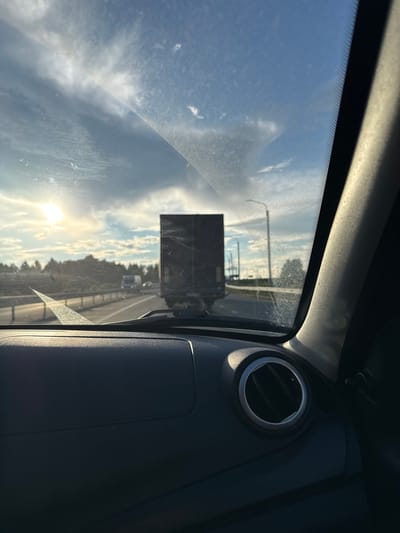 The image is a point-of-view shot from inside a vehicle traveling on a highway in Nagornaya, Russia. The sun is low in the sky, casting a bright light and creating lens flare. The sky is blue with scattered white clouds. In front of the vehicle, a large dark-colored truck is traveling in the same direction, partially obscuring the view of the road and the trees on the side. The dashboard of the car is visible in the foreground, with a circular air vent on the right. The windshield has some streaks and smudges. The overall scene suggests a highway journey during daylight hours.
