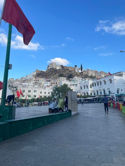 The image depicts a panoramic view of a public square in Moulay Idriss Zerhoun, Morocco, under a clear blue sky with scattered white clouds.
In the foreground, a tall green pole stands on the far left, from which a red Moroccan flag with a green star is partially visible, fluttering to the right. A dark green painted concrete planter or low wall extends horizontally across the lower left portion of the frame. A person in dark attire is bending over this wall on the far left.
The central and midground areas are dominated by a paved pedestrian square composed of grey rectangular flagstones. On the left side of the square, an area features multiple smaller red flags mounted on poles and several white parasols or awnings, beneath which people are seated at tables. Adjacent to the green wall, towards the center-left, a group of three individuals is positioned next to a light-colored concrete block structure. One person is seated on a small wooden stool, facing right, wearing a light-colored top. Another person stands facing the seated individual, dressed in a lime-green robe over darker clothing. A third person, mostly obscured, stands to the right, wearing a blue and yellow patterned garment.
To the right of the paved square, rows of brightly colored plastic chairs (red, yellow, green, blue) are arranged, indicating outdoor seating for a commercial establishment. A person wearing a dark blue shirt and dark pants is walking away from the camera, towards the background, in the middle of the paved area. A street lamp fixture is visible in the upper right.
The background is dominated by a steep hillside densely covered with white, multi-story buildings, characteristic of a traditional Moroccan town. The structures on the upper slopes appear older and more fortified. Sparse green vegetation is interspersed among the buildings on the hillside. Another small red flag is visible on a rooftop in the mid-background to the right of the hillside. The scene is illuminated by natural daylight, casting subtle shadows.