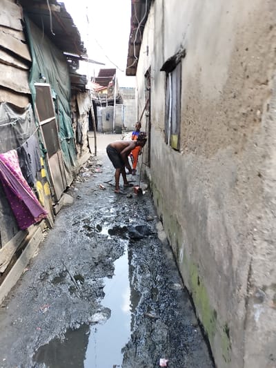 The image depicts a narrow, unpaved alleyway in Oworonshoki, Nigeria, characterized by significant mud, puddles of dark standing water, and scattered refuse. On the left, a structure composed of wooden planks and green tarpaulins supports hanging laundry, including a purple patterned fabric and dark garments. A metal screen panel is also affixed to this structure. On the right, a weathered concrete wall exhibits patches of green moss or algae near its base and features a dark, recessed window opening, from which a dark object protrudes.
In the mid-ground, a shirtless adult male with dark skin and braided hair, wearing dark shorts and flip-flops, is bent forward, facing away from the camera, looking down at the ground near the concrete wall. Adjacent to his feet is a small, portable machine, possibly a generator or pump, with a visible red pull-start handle, connected by a black cable or hose extending towards the wall. Further down the alley, behind the first individual, a second person, possibly a child, wears an orange top with blue shoulders and sleeves. This individual holds a long, thin stick or pipe-like object directed towards the concrete wall. Exposed electrical wiring is visible strung between the structures overhead. The scene is illuminated by daylight from above.