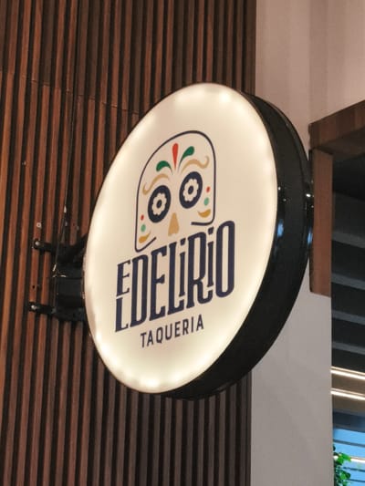 A close-up shot depicts a circular, illuminated sign mounted on a wall with vertical wood slats. The sign is white with a black rim and features a stylized skull design at the top, decorated with red, green, and yellow elements. Below the skull, the name "EDELIRIO" is printed in large, navy blue letters, followed by "TAQUERIA" in smaller, navy blue font. The sign is affixed to the wall by a black metal bracket. Parts of a doorframe and a window can be seen behind the sign. The sign's presence suggests it is the location in Guatemala City, Guatemala.