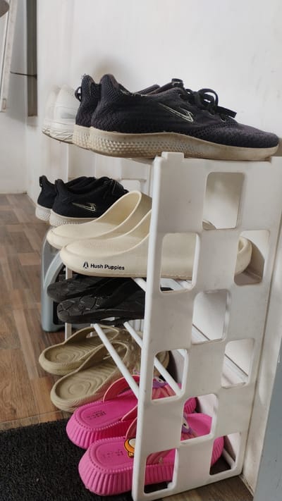 A white plastic shoe rack stands against a light-colored wall, housing various footwear. The top shelf displays two pairs of dark-colored athletic shoes and one pair of white athletic shoes. Below, a pair of cream-colored clogs with "Hush Puppies" branding is visible. The third shelf holds a pair of dark-colored sandals. The fourth shelf contains two pairs of beige flip-flops. The bottom shelf showcases two pairs of bright pink children's slides. A dark grey floor mat is partially visible in the foreground. The scene is likely indoors, possibly in a residence in Banjarsari, Indonesia, given the context.