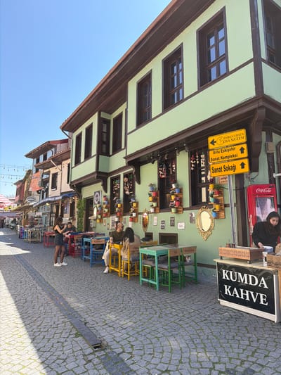 This photograph captures a vibrant street scene in Eskisehir, Turkey, under a clear blue sky. The main subject is a traditional building with light green walls and dark brown wooden beams, featuring numerous windows. Below this building, a lively outdoor cafe or market area unfolds on a cobblestone street.
Several people are present, including a woman standing behind a counter with a red Coca-Cola refrigerator, and others seated at colorful outdoor tables and chairs. One notable subject is a cat comfortably perched on a green chair. The street is paved with gray cobblestones, showing patterns of sunlight and shadow, indicative of a bright, sunny day.
The building walls are adorned with various decorative items, including small potted plants, ornamental mirrors, and framed pictures, creating an inviting and artistic atmosphere. Outdoor furniture, including tables and chairs in green, blue, yellow, and red, are arranged on the sidewalk.
Visible text includes a large black sign displaying "KUMDA KAHVE" (Sand Coffee). Yellow directional signs point to "IMREN ERSEN OYA MÜZESI" (İmren Erşen Embroidery Museum), "Avlu Eskişehir Sanat Kompleksi" (Courtyard Eskişehir Art Complex), and "Sanat Sokağı" (Art Street). A smaller sign indicates "ARİFBEY SOKAK". On the backs of the chairs, words such as "Sadakat" (Loyalty), "Aşk" (Love), "Mutluluk" (Happiness), and "Ölüm" (Death) are inscribed. In the background, signs for "SÜRMELİ KONAK" and "inci börek" are also visible. The overall setting suggests a bustling yet relaxed cultural and commercial street.