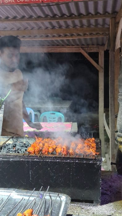 Skewered meat is being grilled over hot coals, generating a dense cloud of smoke. The food stall appears to be constructed with corrugated metal and timber framing. A man stands near the grill, seemingly tending to the cooking process. Behind him, blue plastic chairs are visible. A metal tray filled with skewers sits to the right of the grill.