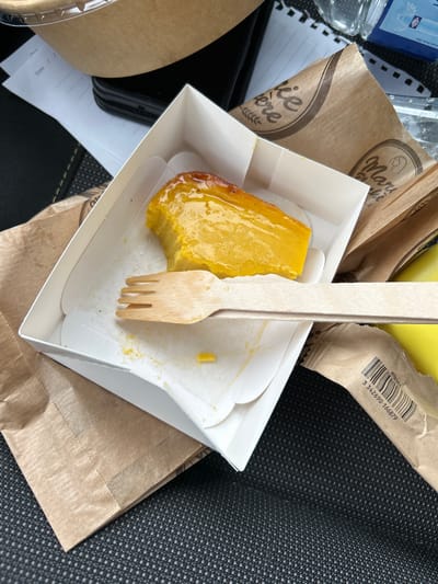 In an indeterminate indoor location, perhaps a car, an eaten pastry from Marie Blachère is the focal point. The pastry, golden brown and partly consumed, sits in a white cardboard container with a scalloped edge. A wooden fork is stuck into the pastry. The container is placed on a piece of brown paper and is surrounded by wrappers from Marie Blachère and a KITBOIS wrapper with a barcode visible, plus a bottle of water. There is also a form that has date fields on it. Based on the Marie Blachère wrappers, the location is presumed to be in or near Valence, France.