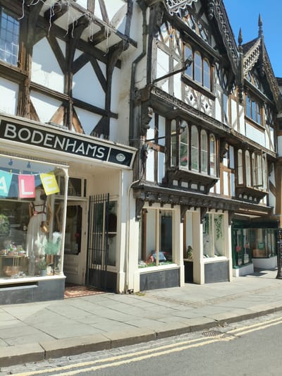 The image shows the exterior of a shop named "BODENHAMS" in Ludlow, United Kingdom. The building features traditional timber-framed architecture with white infill and dark wooden beams. The shop has large display windows showcasing merchandise, including mannequins. Signs indicating a sale are visible in one of the windows. The street in front of the shop is paved with stone slabs, and a road with yellow lines is visible in the foreground. The sky is clear and blue. The image appears to be taken on a sunny day.