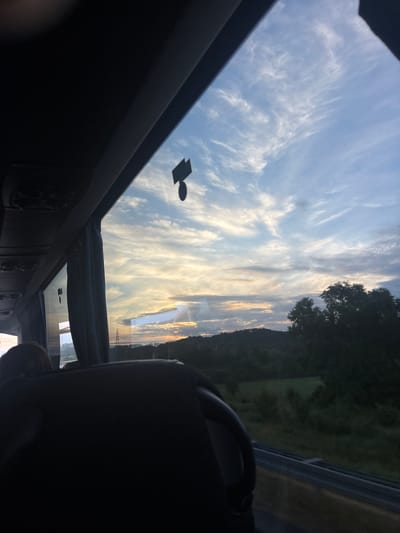 The image is an interior view of a vehicle, possibly a bus or van, looking through a sunroof. Above, the sky is visible, filled with wispy, cirrus-like clouds. Outside the vehicle, a landscape is partially visible, with greenery suggesting trees or hills. Power lines are seen through the sunroof. A small, round sticker with a rectangular addition is on the sunroof. Inside the vehicle, elements of the interior, such as seating and dashboard details, are seen. The image appears to be taken while the vehicle is in motion. The location is Obrenovac Urban Municipality, Serbia, deduced from the style of power lines and landscape.