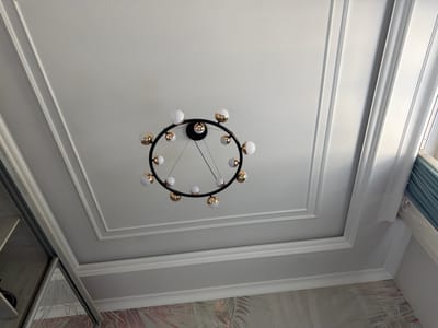 The image shows a circular chandelier with multiple white and gold globe lights hanging from a white, multi-tiered coffered ceiling. The chandelier is centrally located in the frame. The ceiling has ornate molding details. To the left, a portion of a wardrobe with a mirrored door is visible, reflecting a wall with tropical-themed wallpaper. To the right, light blue curtains are partially visible near a window. The scene appears to be an interior residential setting. There are no people or animals present in the image. The location is not specified as Джапалак, Kyrgyzstan.