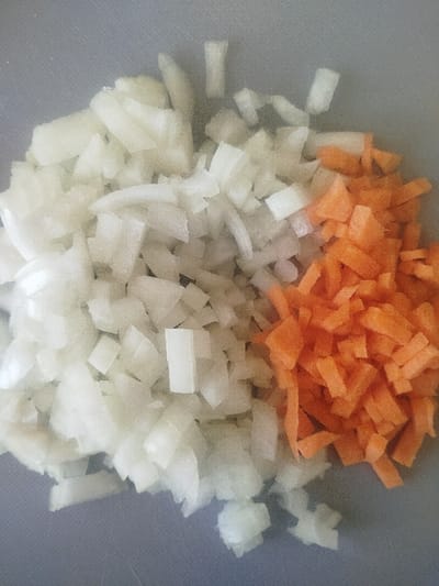 In the city of Klenovik, Bulgaria, a close-up overhead view captures two distinct piles of diced vegetables resting on a light gray, textured surface, likely a cutting board. The larger pile, occupying approximately two-thirds of the frame on the left, consists of numerous irregularly cut, translucent white pieces, consistent with diced onion. The adjacent pile, positioned on the right and slightly smaller, comprises numerous small, uniformly diced, vibrant orange pieces, consistent with chopped carrot. The vegetables are arranged separately, indicating preparation for a culinary purpose. No individuals or additional objects are visible.
