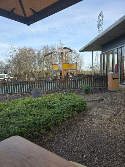 This image depicts an outdoor recreational area and adjacent building in Urvillers, France, under a clear, bright sky. In the foreground, the edge of a light-colored wooden surface, possibly a table or bench, occupies the lower left. Adjacent to it on the left is a dense, manicured patch of green shrubbery, behind which a dark grey potted plant is visible. The ground is covered with a dark, speckled gravel surface.
A brown wooden picket fence forms a barrier across the mid-ground. Beyond this fence, a children's playground structure constructed from light-colored wood, featuring yellow and grey panels and a slide, is positioned centrally. A white, bird-shaped weather vane, resembling a goose, is mounted at the apex of the playground structure.
To the right of the playground, the exterior of a modern building with large, reflective glass windows and a prominent, flat grey concrete roof overhang is visible. A wooden trash receptacle with a dark vertical stripe is situated near the building's facade. In the background, a dense line of leafless deciduous trees stretches across the horizon. A white van is partially visible behind these trees on the far left. A tall, metallic lattice-type communications tower rises prominently into the sky behind the building on the right. Faint, parallel overhead lines are discernible across the blue sky. No individuals are present in the scene.