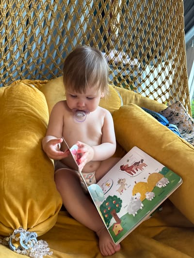 A young child is sitting in a yellow cushioned hammock chair, reading a book. The child is light-skinned, with fair hair and a pacifier in their mouth, and is wearing a diaper. The book is open to a page featuring illustrations of animals and plants, with Russian text visible. The hammock chair has a woven net structure. Additional pillows with floral patterns are in the background. The location is unspecified but is unlikely to be Чёрное, Russia, due to the language on the book being Russian, not exclusively associated with that city.
