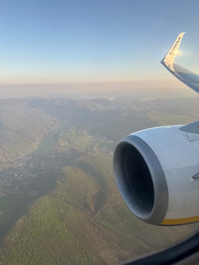 The image shows an aerial view from a Ryanair airplane. A portion of the wing with the Ryanair logo is visible in the upper left corner. A white engine is prominently displayed on the right side of the image. Below, the landscape is visible, including fields, forests, a river, and a town possibly obscured by haze. It is likely that the plane is flying over Hungary, more specifically the city of Vecsés.