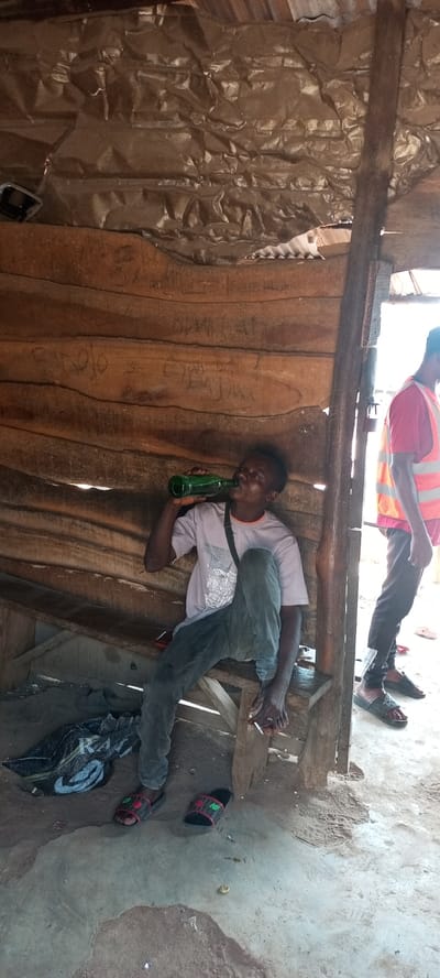 This image captures an informal scene in Iyara, Nigeria, featuring two men within and beside a rustic wooden structure.
The primary subject is a young man, seated on a low wooden bench inside the structure. He wears a light-colored t-shirt with a graphic design, dark trousers, and a black shoulder bag. He is actively drinking from a green glass bottle, tilting his head back to do so. In his right hand, he holds a lit cigarette between his fingers. His bare feet are visible, with a pair of red and black slides (one bearing an "NB" logo) discarded on the dirt and concrete floor near his feet. A black bag or sachet with "RAV" and a white logo printed on it lies on the ground next to him.
To the right, partially visible at the entrance of the structure, stands another man. He is wearing a red t-shirt underneath a bright orange high-visibility vest, dark trousers, and black slides. His face is not visible, and he appears to be standing casually.
The setting is a simple, possibly makeshift shelter constructed from rough-hewn wooden planks that form the walls and a low bench. The ceiling appears to be made of crumpled corrugated metal or tin sheeting. Inscriptions, possibly names or words, are scratched into the wooden wall behind the seated man, including faintly visible words like "BINDOLO" and "BENJUI." A small padlock hangs on the wooden frame near the standing man. The ground inside is a mix of dirt and uneven concrete, with a small white cap or lid also visible on the floor.
The scene is illuminated by bright natural daylight, suggesting it is daytime. The overall atmosphere is casual and informal.