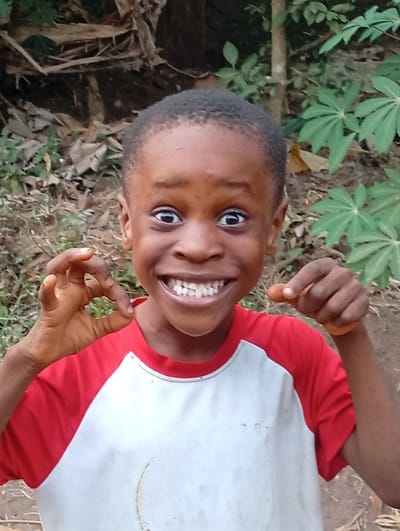 A young boy with short, dark hair smiles broadly, showcasing his teeth. He wears a white t-shirt with red sleeves. His eyes are wide and expressive, and he is making a gesture with both hands, touching his thumb and forefinger together. The background is a slightly blurred outdoor scene, with greenery including various plants and trees, suggesting a natural or rural setting.