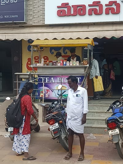 The image shows a street-level view of a shop with the name "MANASA" in red Telugu script on a white background above a yellow awning. A sign with a phone number "9091336699" is visible on the left. In front of the shop, a food stall or tea counter is set up. Several people are present. A woman in a red and white patterned top and white patterned pants is standing on the left, wearing a black backpack. Next to her is a scooter. A man in a white shirt and dhoti is standing in the foreground, looking to his left. Behind him, other people are seen near the shop entrance. Motorcycles are parked in the foreground and on the right. The ground is paved. The overall scene appears to be a typical street in Kurnool, India.
