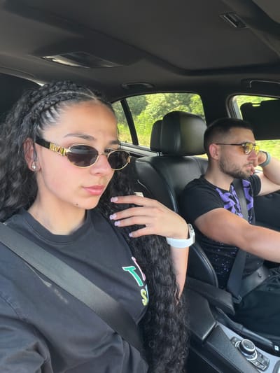 The media file shows two people in the front seats of a car. The person in the foreground is a woman with dark curly hair, wearing sunglasses and a black t-shirt. She has braids on the side of her head and a white smartwatch on her left wrist. The person in the background is a man with short dark hair and a beard, also wearing sunglasses and a black t-shirt with a purple design. He is leaning his head back, with his arms crossed. The scene appears to be during the daytime, with sunlight filtering through the car windows and trees visible outside. The overall mood is relaxed and casual. There is no visible text in the image. The location context provided is Khadzhiysko, Bulgaria.