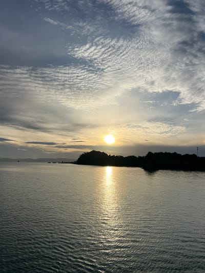 The image depicts a serene sunset over the Gulf of Thailand, likely near Tambon Don Sak in Thailand. The sun, a bright orb, is positioned low in the sky, casting a shimmering golden reflection on the calm water. Wispy, textured clouds, characteristic of cirrocumulus formations, stretch across the sky, tinged with hues of orange and yellow from the setting sun. In the foreground, the water is rippled, creating a textured surface that reflects the light. Silhouetted against the bright sky is a dark, tree-lined island or peninsula, with the faint outlines of structures and a tall antenna visible on its edge. In the distance, hazy mountains form a soft backdrop, and a few small boats are visible on the horizon. The overall scene is one of natural beauty and tranquility, capturing the peaceful ambiance of a coastal sunset.