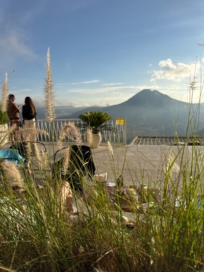 The image shows a landscape in Kedisan, Indonesia, featuring a mountain in the background under a partly cloudy sky. In the foreground, there is tall grass with seed heads obscuring some of the scene. A paved area with a railing is visible, and several people are present, though partially obscured by the grass. There is a potted plant and a yellow sign on the paved area. People in the scene are engaged in various activities, including sitting and looking at the landscape.