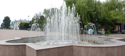 A wide-angle, eye-level shot captures a public plaza with a prominent fountain in the foreground. The fountain, constructed from stone, is actively spraying water upwards in multiple streams, creating a dynamic and slightly blurred effect. Inscribed graffiti is visible on the fountain's surface.
In the mid-ground, several people are present. To the right of the fountain, a young girl with blonde hair, wearing a light green jacket and pants, is holding a paper or object towards the fountain. To the left, another individual, seen from behind, wears a light blue backpack and pink pants, facing away from the camera. Further back, a child in a blue shirt and black shorts stands near the edge of the fountain, partially obscured by water. A black metal fence runs along the back of the plaza.
The background features a dense line of tall, mature trees with lush green foliage, characteristic of willow trees. Beyond the trees, a multi-story building with a light-colored facade, red-tiled roof, and numerous windows is visible, suggesting an urban or town setting. The sky is overcast and appears bright, indicating a cloudy day. The ground in the plaza is paved with interlocking stones. The overall scene appears to be located in a park or public square within the city of Kirsanov, Russia, given the presence of the fountain, trees, and architecture.