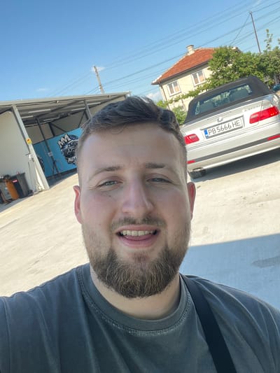 A selfie photograph taken during the day shows a man with a light beard wearing a gray T-shirt with a black strap over his shoulder. The license plate PB 5666 HE of a silver car is visible in the background, along with a building with a red tiled roof. There's also a partially obscured banner with "AUTO" printed on it. The location is Sadovo, Bulgaria.