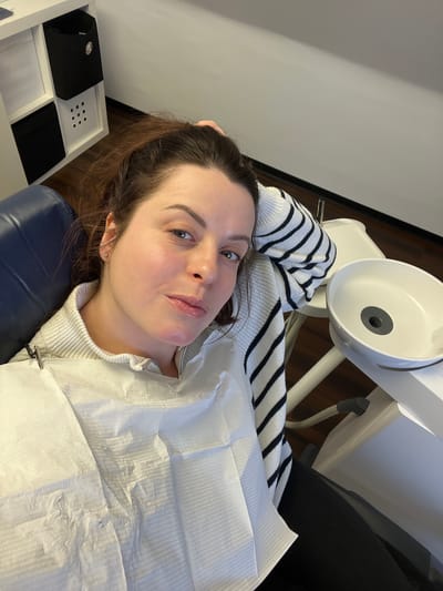 An adult female subject is observed reclining in a blue dental examination chair within a clinical setting in Monsheim, Germany. Her gaze is directed towards the camera. The subject presents with fair skin, light-colored eyes, and reddish-brown hair, partially secured. She is dressed in a light-colored ribbed sweater, beneath a white disposable dental bib fastened by a metal clip at her throat. A white sweater sleeve with black horizontal stripes is visible on her left arm, which is positioned behind her head. A subtle reddish discoloration is apparent on her chin/jawline. Adjacent to the subject's right, a white ceramic dental cuspidor with a dark grey drain is mounted on a white medical apparatus. In the background, to the left, a white modular shelving unit contains dark fabric storage bins. The environment features light-colored walls and dark wooden flooring.