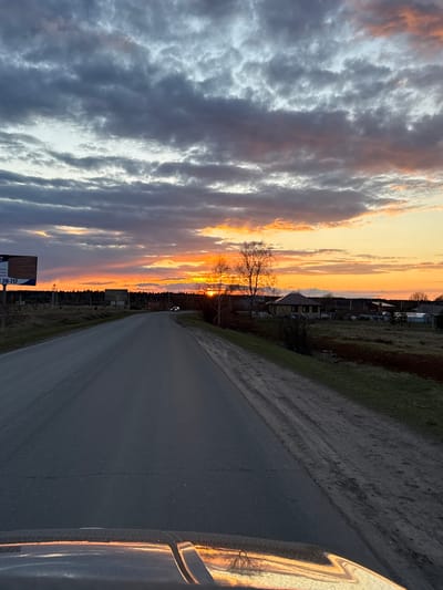 The image, taken in Vorob'evo, Russia, captures a dramatic sunset over a rural landscape. The setting sun, a brilliant orb of orange and yellow, is positioned on the horizon, casting a warm glow across the sky. Dark, textured clouds dominate the upper and left portions of the frame, showing shades of grey, purple, and hints of orange where the sunlight catches them.

A narrow, unpaved or dirt road stretches into the distance, flanked by grassy verges. On the left side of the road, a prominent bare tree stands silhouetted against the vibrant sunset colors. Further along the horizon, several distant houses or buildings are visible among dark trees.

In the immediate foreground on the right, the polished surface of what appears to be a car's side mirror or window frame reflects the brilliant sky and the tree. On the left side of the road, a dark, rectangular sign is partially visible. The numbers "20-717" are clearly legible on the sign, along with partial words or letters such as "mm" and "ome."

The scene depicts the tranquil end of a day, likely viewed from a moving or parked vehicle, with the dramatic cloudscape and sunset serving as the primary subjects. The bare tree suggests a late autumn, winter, or early spring season.