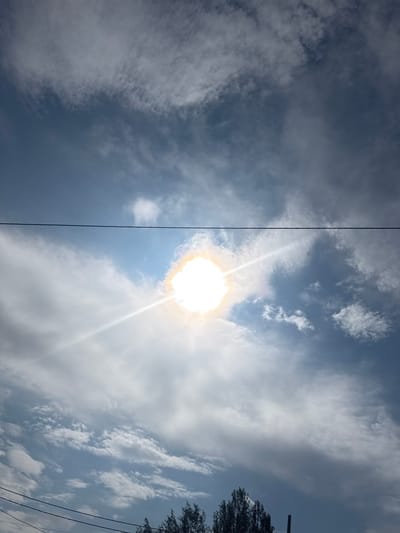 This media file shows a bright sun shining through a cloudy sky. The sun is positioned in the center-right of the frame, emitting strong rays and creating a halo effect. A thin, dark vertical line runs down the left side of the image, possibly a power line or a thin pole. In the upper right corner, the dark silhouette of tree branches and two parallel power lines are visible against the sky. The sky itself is a mix of dark and light clouds, with patches of clear blue visible. The scene suggests a daytime setting, with the bright sun indicating fair weather, though the clouds suggest some atmospheric activity. The presence of power lines and trees hints at an outdoor, possibly semi-urban or rural environment. No people or text are visible in the media file.