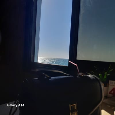 From inside a building in Al-Mawasi, the sea is visible through a window. A pink straw sticks out from a dark-colored object next to the window. Other objects, including a green plant and a paper labeled "SKY RESTAURANT", are also visible.