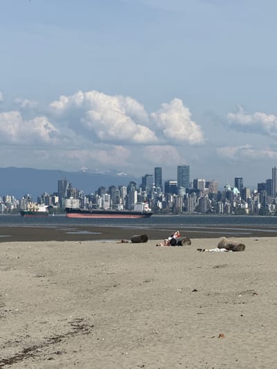 The image depicts a beach scene in Electoral Area A, Canada, facing the Vancouver skyline across the water. The sky is light blue with large, puffy white clouds. The beach is sandy and runs along the right side of the frame. A cargo ship is visible in the water, positioned between the shoreline and the city. A few people are seated on logs.